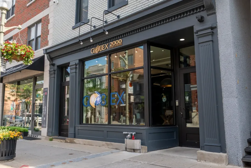 Angled view of the storefront cornice, gooseneck lamps, and logo-marked display windows beneath a white brick facade