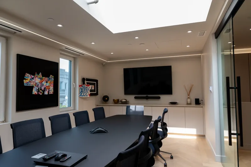 Boardroom beneath a skylight, with a long dark table, mesh chairs, and a wall-mounted screen