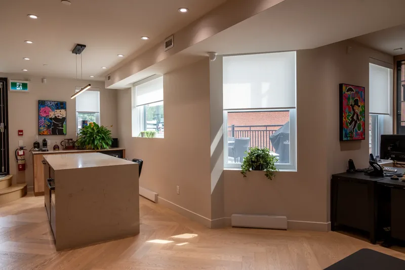 Kitchen island and work area beside tall windows with roller shades and colourful artwork
