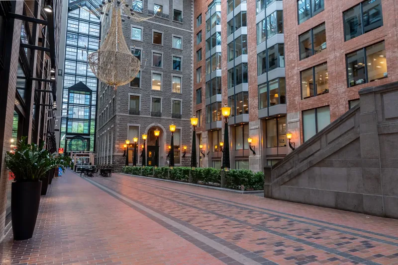 Brick-paved atrium promenade with lanterns, planters, and a glass roof above stone and brick facades