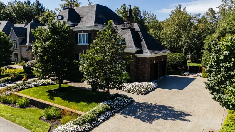 Paved driveway bordered by layered garden beds and mature trees