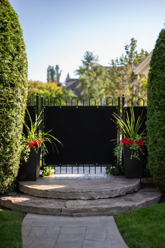 Black garden gate with planters and curved stone steps between tall cedar hedges