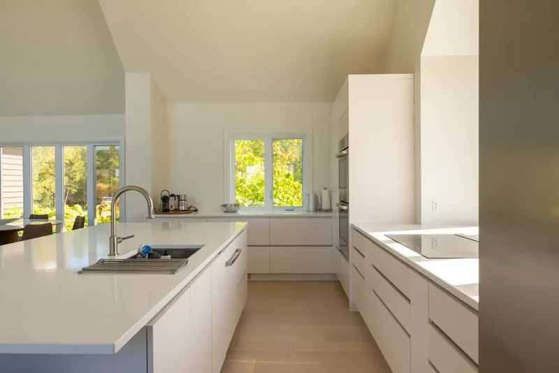 White kitchen with long counters, an island sink, and windows to the trees