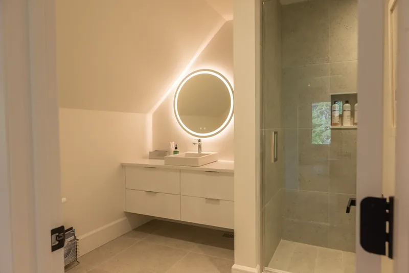 Floating vanity with a round backlit mirror beneath a sloped ceiling