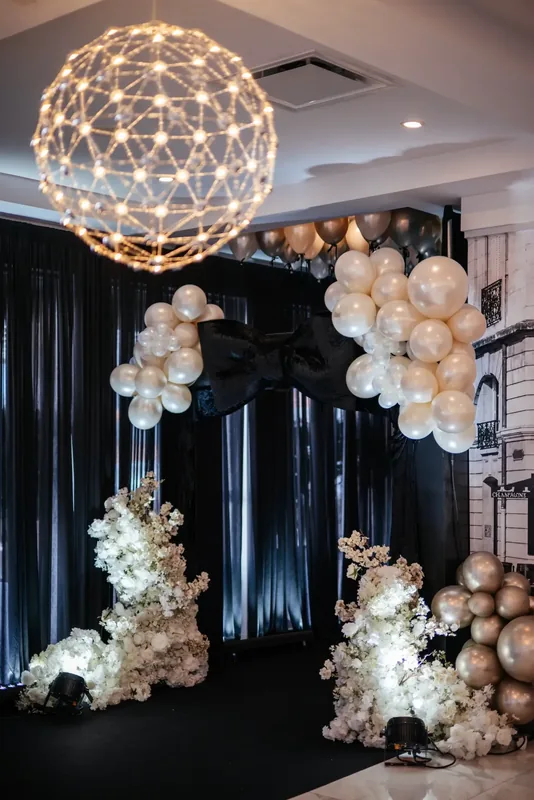 Event backdrop with a lit geometric pendant, pearl balloons, white florals, and black drapes