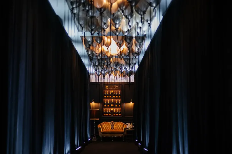 Black velvet corridor under a ceiling of silver balloons, leading to a gold tufted settee and a lit champagne display