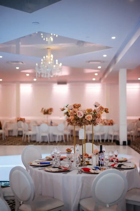 Round table with blush roses on a gold stand and gold chargers, beneath a chandelier near the white dance floor