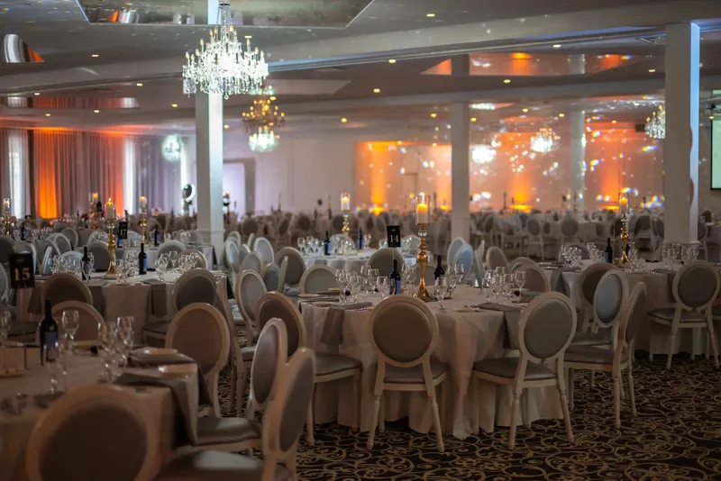 Large hall filled with round tables, gold candle holders, crystal chandeliers, and warm amber uplighting