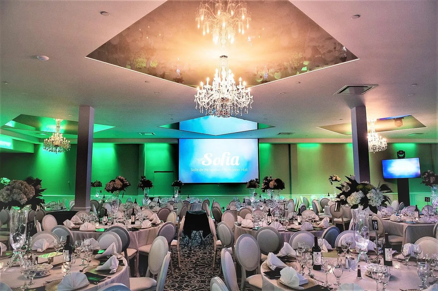Main reception hall with crystal chandeliers in mirrored ceiling trays, green uplighting, and a screen showing the Sofia Reception Hall name